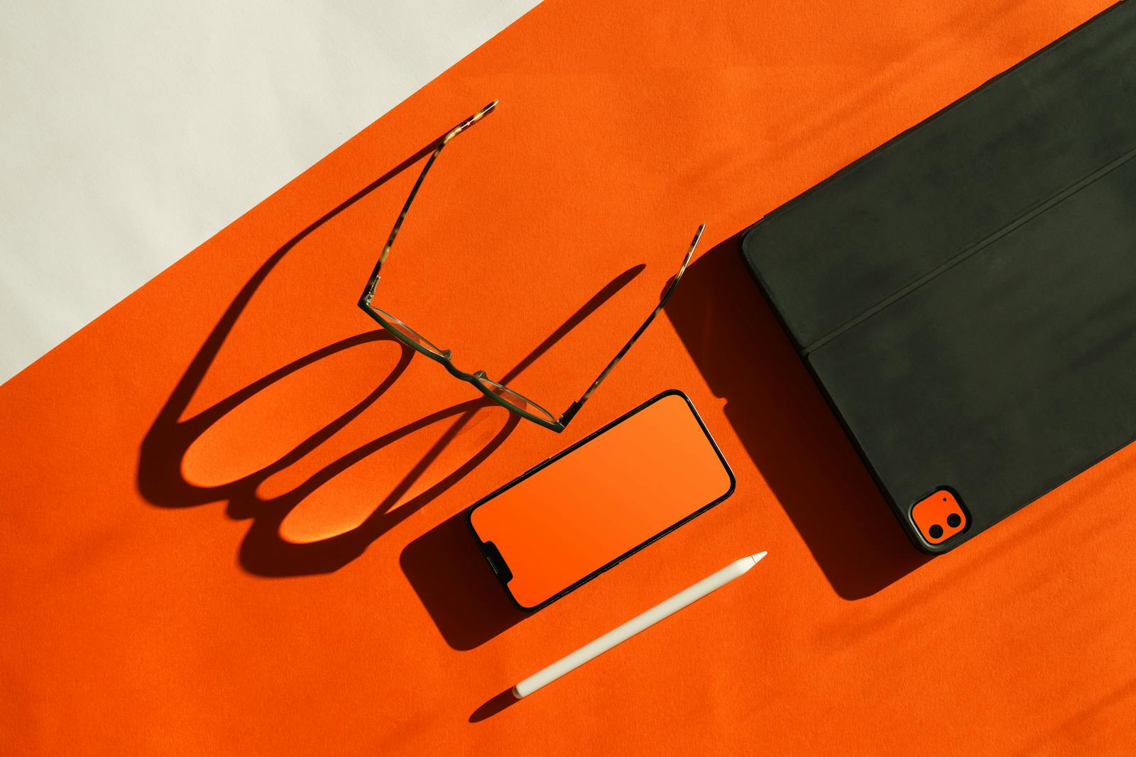 Glasses, smartphone, tablet, and stylus arranged on an orange background with shadows.